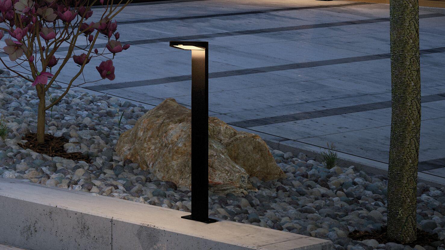 Black metal solar bollard light with warm white illumination in modern garden setting with stones and tree.