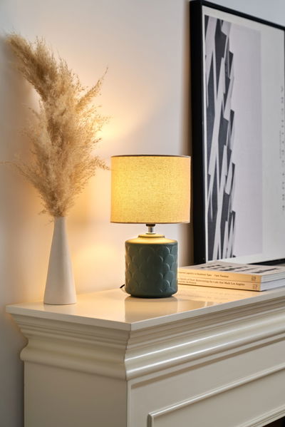 Tischlampe mit grünem Keramikfuß und beigem Stoffschirm auf weißem Kaminsims mit Deko.
