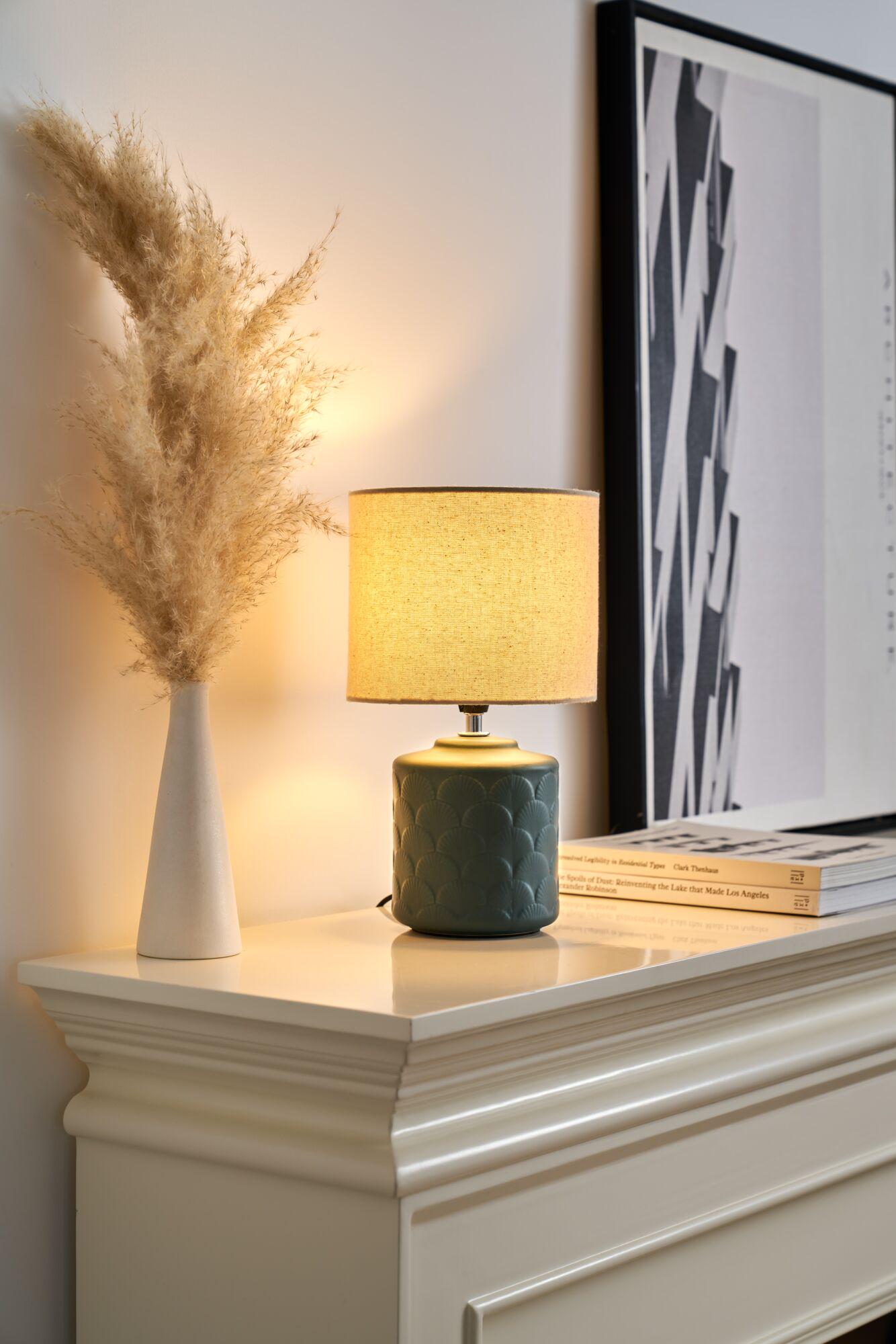 Green ceramic table lamp with beige fabric shade on white mantelpiece beside pampas grass vase