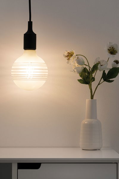 Hangende matte glazen lamp met horizontale strepen naast witte vaas met witte bloemen op tafel