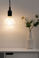 Hangende matte glazen lamp met horizontale strepen naast witte vaas met witte bloemen op tafel