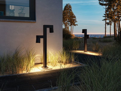 Black modern outdoor metal wall light with warm white illumination by house wall and sandy path.