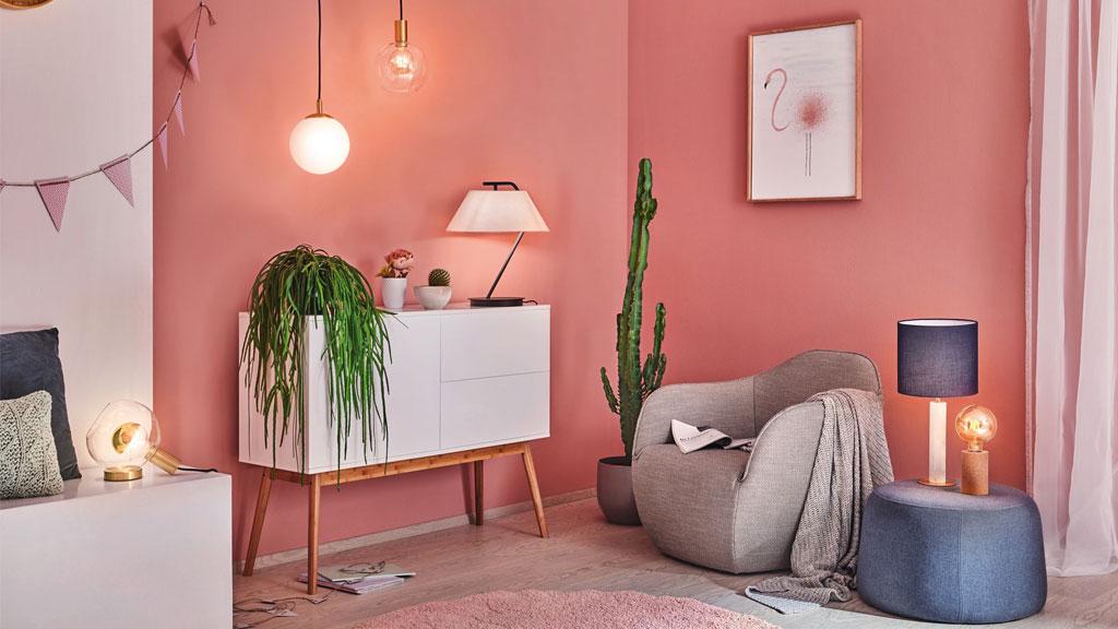Modernes Wohnzimmer mit rosa Wand, grauem Sessel, blauer Lampe und weißen Möbeln für gemütliche Beleuchtung.
