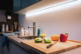 Plan de travail de cuisine moderne avec évier en acier inoxydable, planche à découper en bois et éclairage LED blanc.