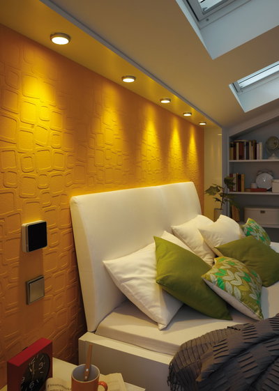 Chambre avec lit rembourré blanc et coussins verts, spots encastrés blanc chaud sur mur orange.