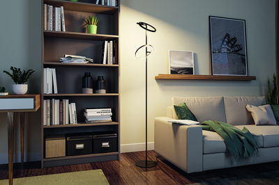 Black LED floor lamp with two circular light rings beside beige sofa in modern living room with bookshelf.