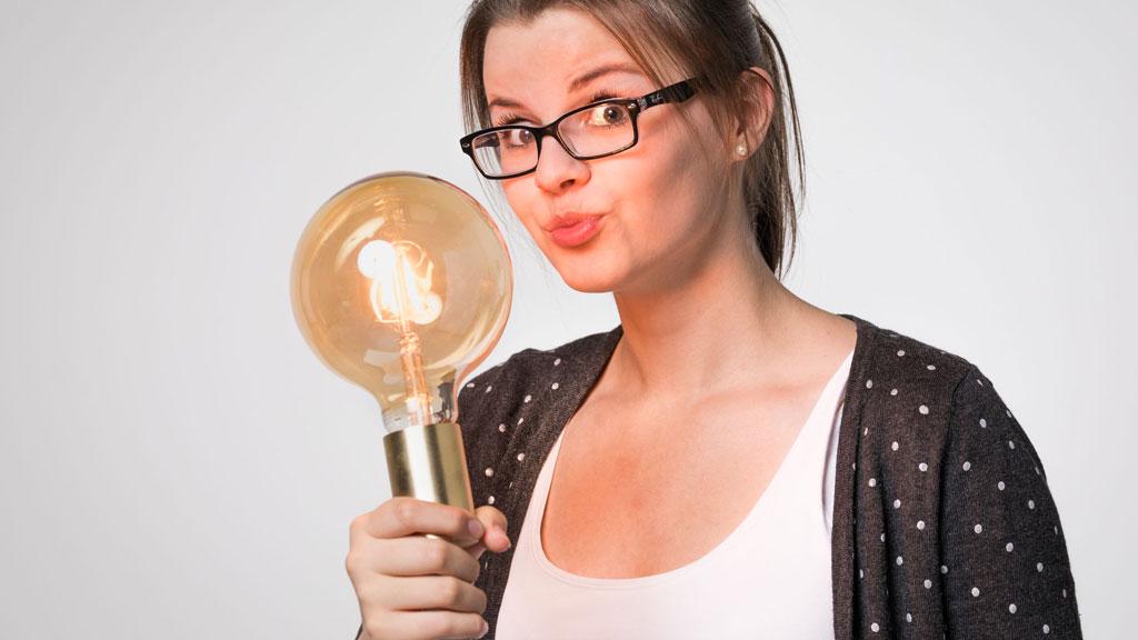 Woman with glasses holding large filament light bulb, symbolizing innovation and lighting technology.