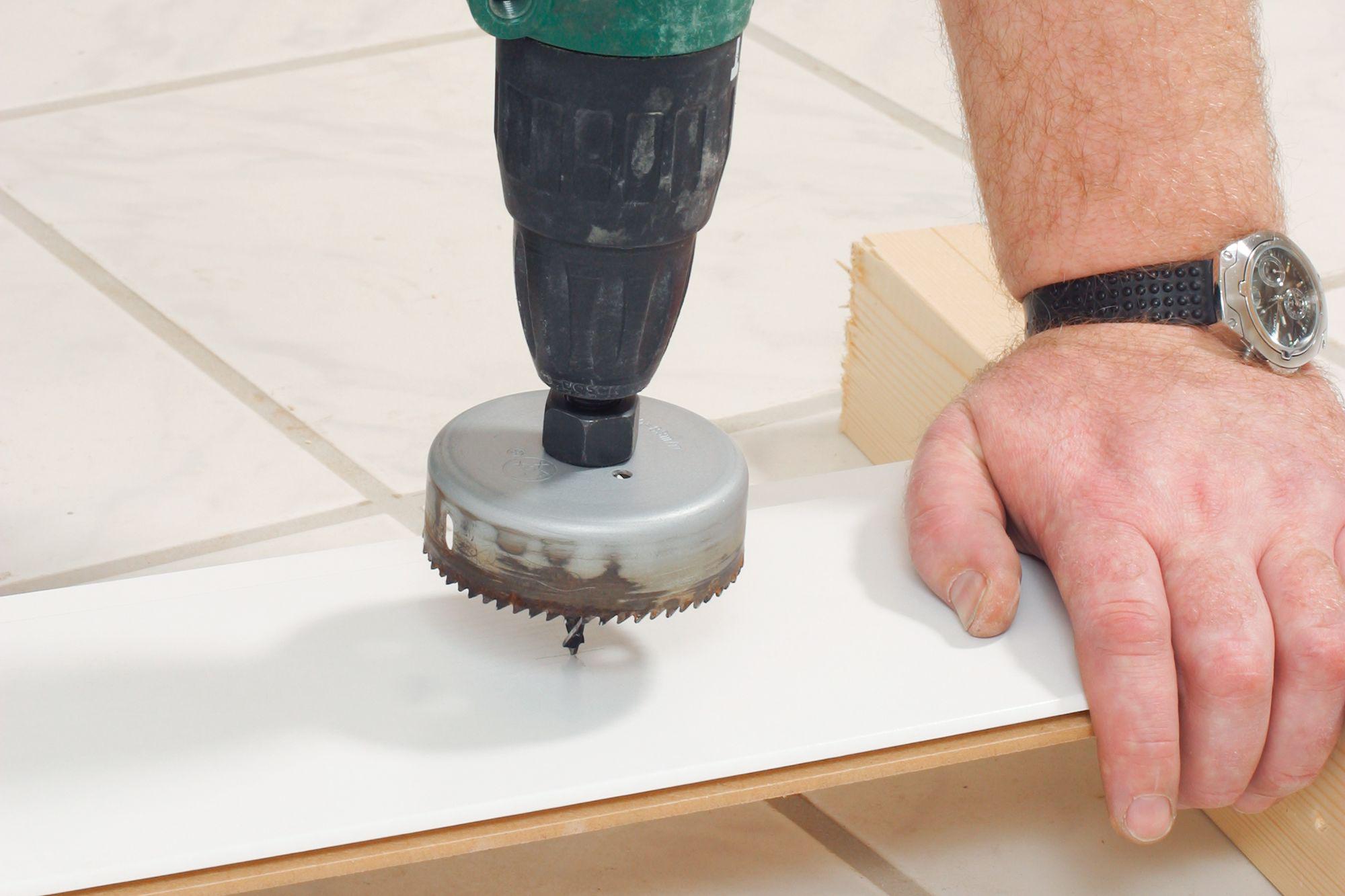 Drill with hole saw cutting white plastic panel on tiled floor, hand stabilizing the workpiece