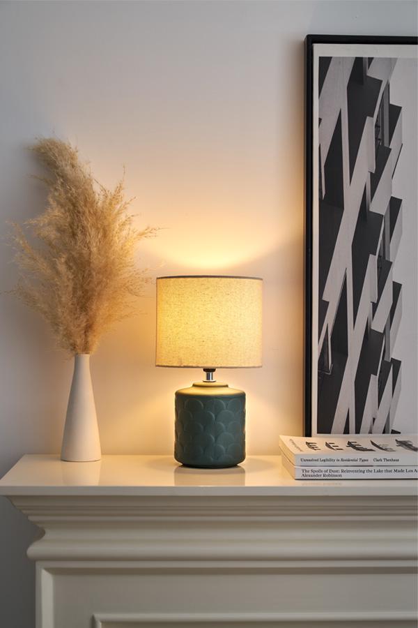 Table lamp with green ceramic base and beige fabric shade on white mantel with decor and artwork.