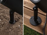 Black metal mount for outdoor spotlight on soil and wooden deck, weatherproof garden lighting fixture.