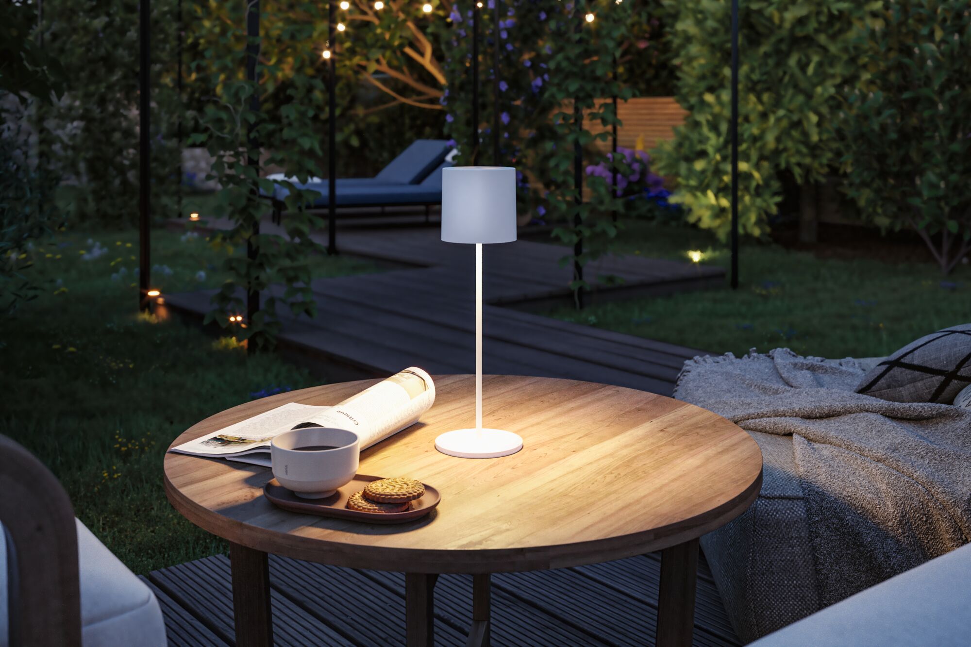 Witte metalen LED-tafellamp op houten tuintafel met kopje, koekjes en krant bij avondlicht