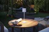 Witte metalen LED-tafellamp op houten tuintafel met kopje, koekjes en krant bij avondlicht