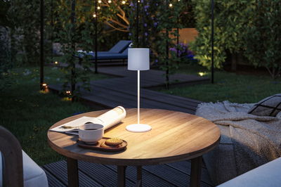 Witte metalen LED-tafellamp op houten tuintafel met kopje, koekjes en krant bij avondlicht