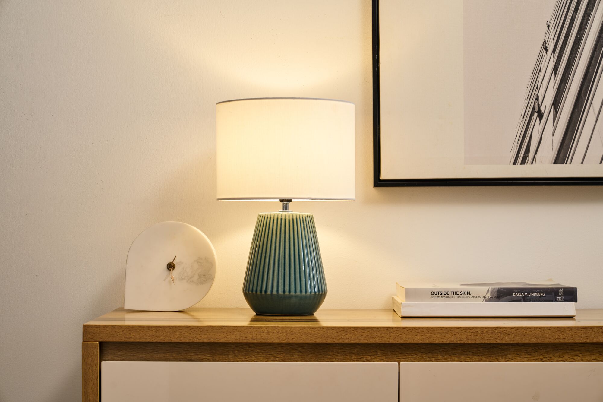Table lamp with green ceramic base and white fabric shade on wooden sideboard with marble clock and books