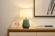 Table lamp with green ceramic base and white fabric shade on wooden sideboard with marble clock and books