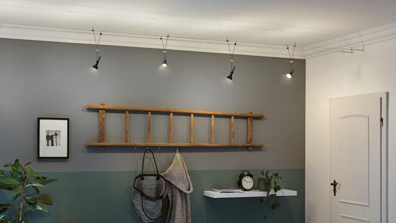 Moderne kelderverlichting met vier zwarte plafondspots aan draaddraden boven een grijze muur met houten ladder.