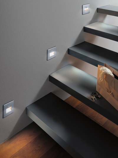 Moderne LED-Treppenbeleuchtung in silbernem Aluminium mit warmweißem Licht an grauer Wand.
