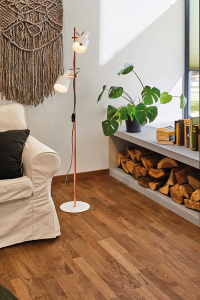 Copper floor lamp with white shades beside beige sofa and stacked firewood in cozy living room.