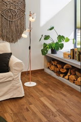 Stehende Kupferlampe mit weißen Lampenschirmen neben beigem Sofa und Holzstapel im Wohnzimmer.