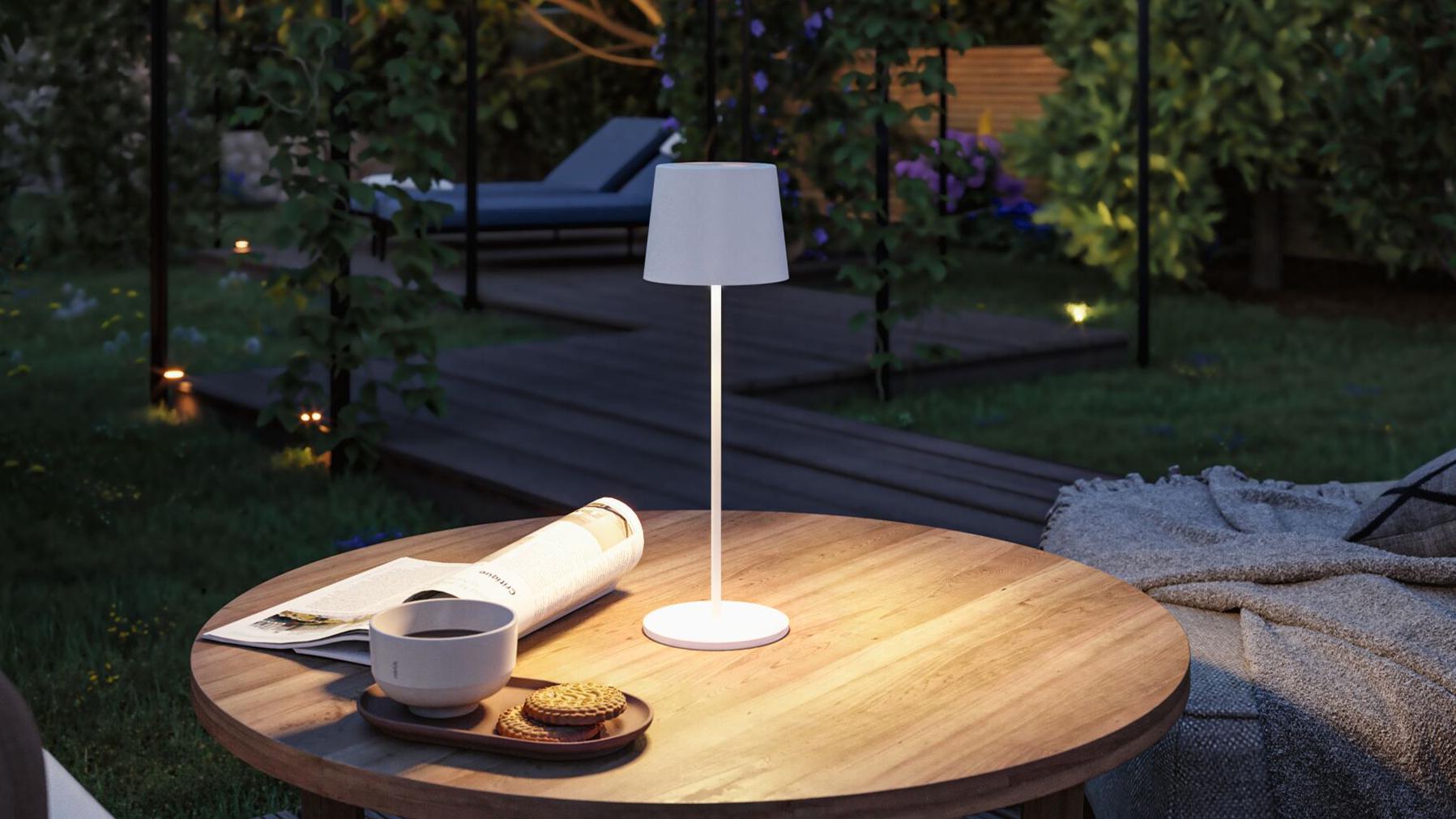 Weiße mobile Akku-Tischleuchte mit Stoffschirm auf Holztisch im Garten mit Keksen und Kaffee.