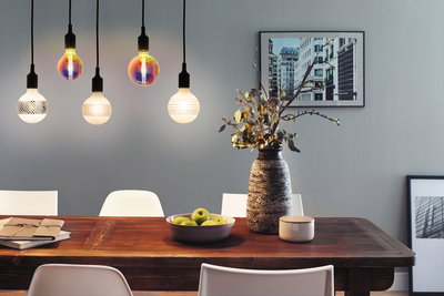 Moderne Pendelleuchten mit bunten und weißen Glühbirnen über Holztisch mit Dekoration und Äpfeln