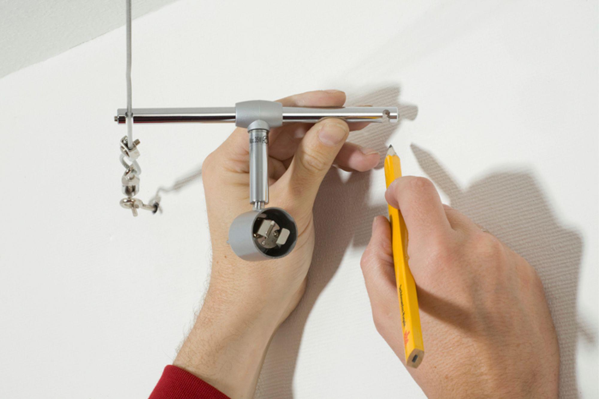Hånd holder sølvfarvet skinnelygte mod hvid væg og markerer borepunkt med gul blyant