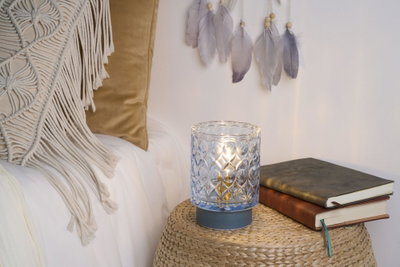 Patterned glass candle holder with blue base on woven table near books and cushions