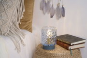 Glazen windlicht met patroon en blauwe voet op gevlochten tafel naast boeken en kussens