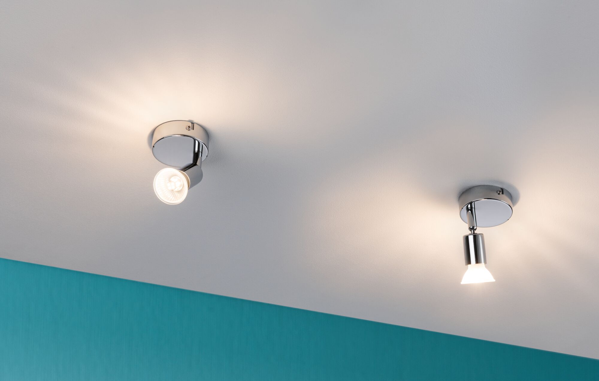 Two chrome LED ceiling spotlights with warm white light on white ceiling and turquoise wall.