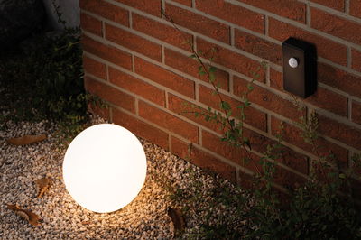 Ronde witte LED-tuinlamp op grind bij rode bakstenen muur met zwarte bewegingssensor.