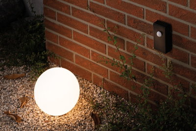 Round white outdoor lamp on gravel by red brick wall with black plastic motion sensor