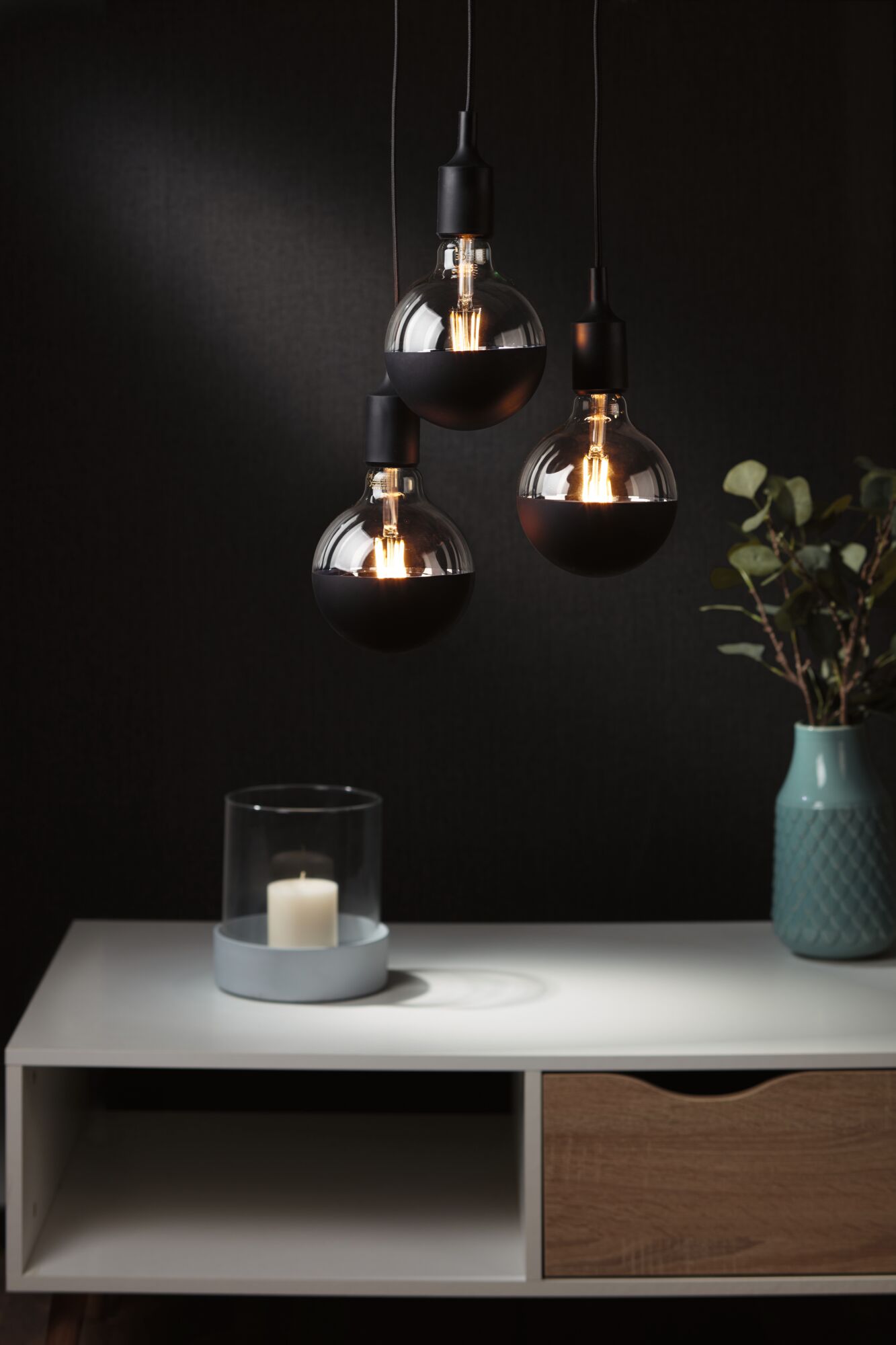Trois ampoules suspendues avec revêtement noir semi-transparent au-dessus d'une table blanche avec bougie et vase
