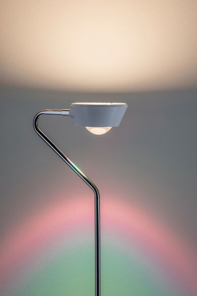 Moderne vloerlamp met witte kap en chromen metalen frame, kleurrijke lichtaccenten op de muur.