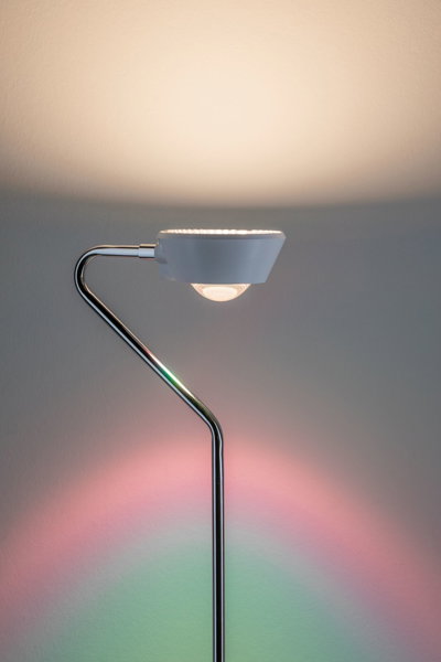 Modern floor lamp with white shade and chrome metal frame, colorful light accents on wall for ambient lighting.