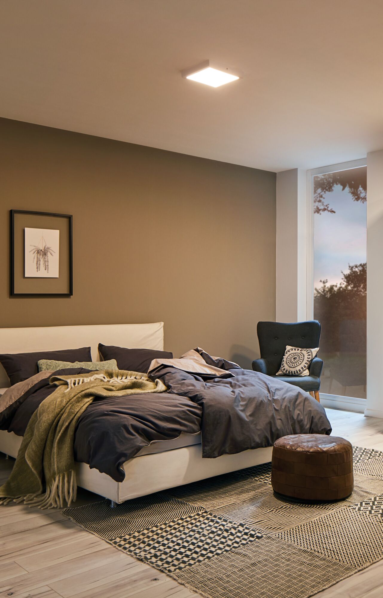 Chambre moderne avec lit beige, literie gris foncé, plaid vert et pouf en cuir marron sur tapis à motifs