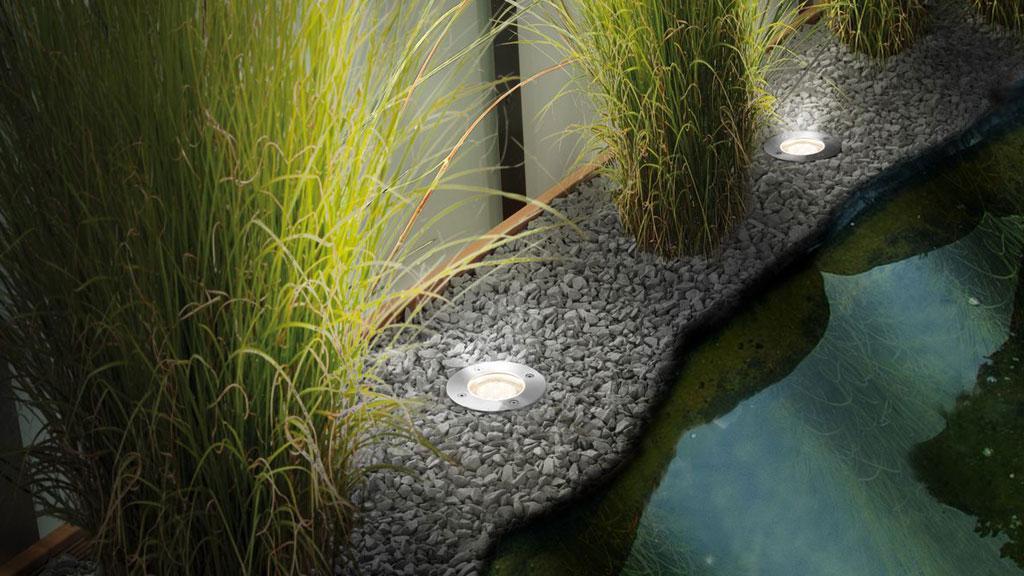 Round white LED pond light in gravel bed by water and grasses for garden lighting.