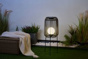 Black metal floor lamp with round light bulb in garden setting with plants and blanket