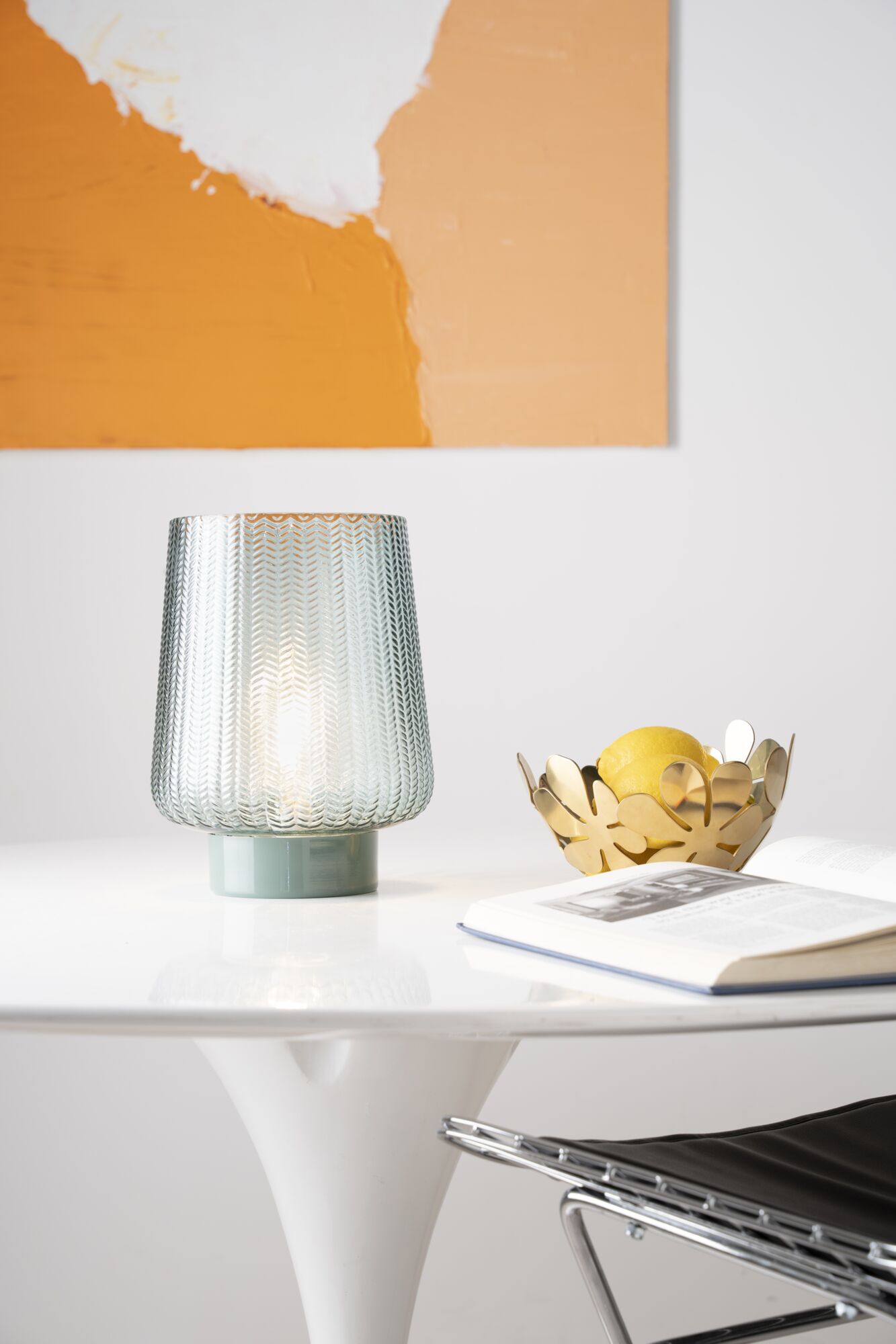 Glas-Tischleuchte mit Muster und grünem Sockel auf weißem Tisch neben goldener Obstschale und Buch