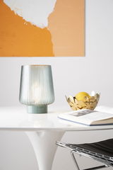 Patterned glass table lamp with green base on white table next to gold fruit bowl and open book