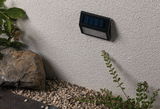 Black plastic solar wall light mounted on white exterior wall with gravel and garden plants