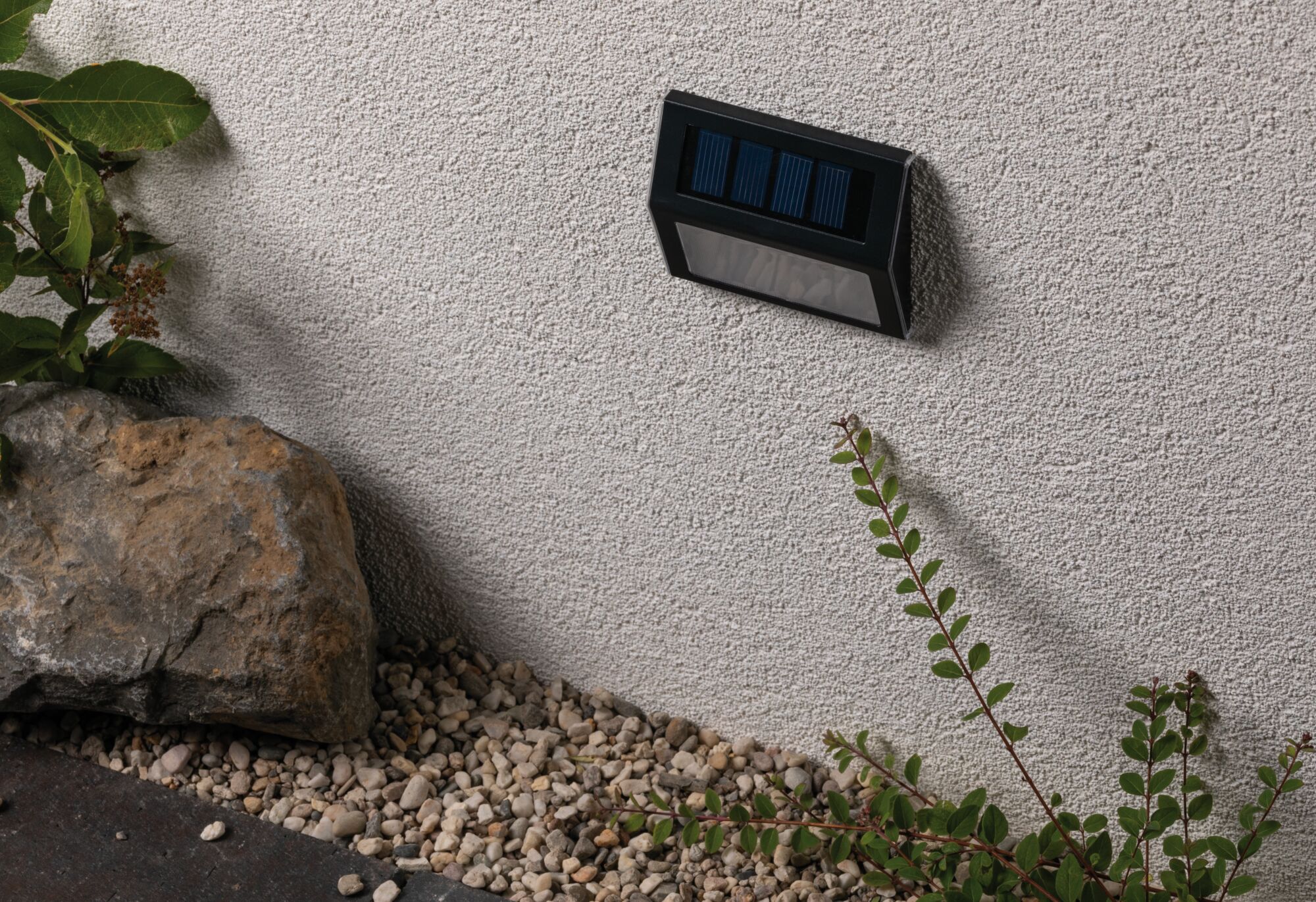 Schwarze Solar-Wandleuchte aus Kunststoff an weißer Außenwand mit Kies und Pflanzen im Garten