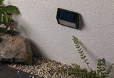 Schwarze Solar-Wandleuchte aus Kunststoff an weißer Außenwand mit Kies und Pflanzen im Garten