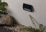 Black plastic solar wall light mounted on white exterior wall with gravel and garden plants