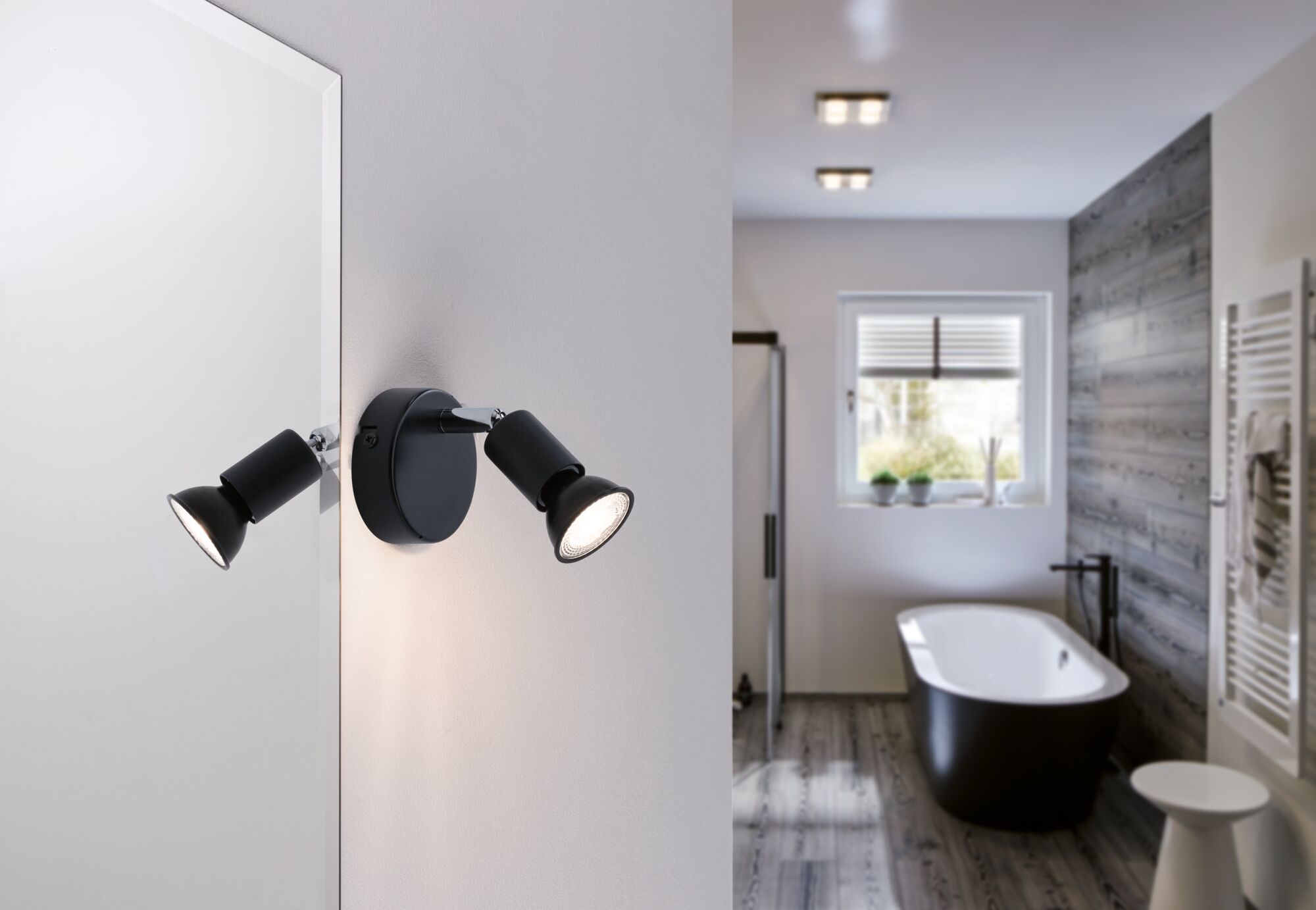 Sort LED-væglampe med to justerbare spots i moderne badeværelse med badekar og trægulv.