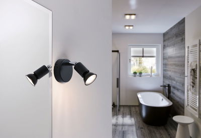 Sort LED-væglampe med to justerbare spots i moderne badeværelse med badekar og trægulv.