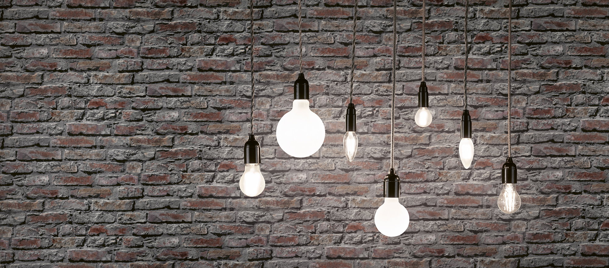 Seven filament light bulbs with black fittings hanging in front of grey brick wall, decorative lighting design.