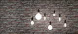 Seven filament light bulbs with black fittings hanging in front of grey brick wall, decorative lighting design.