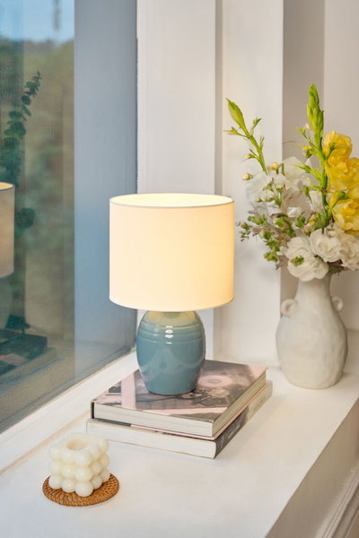 Keramische tafellamp met blauwe voet en witte stoffen kap op boeken bij raam met bloemenvaas.