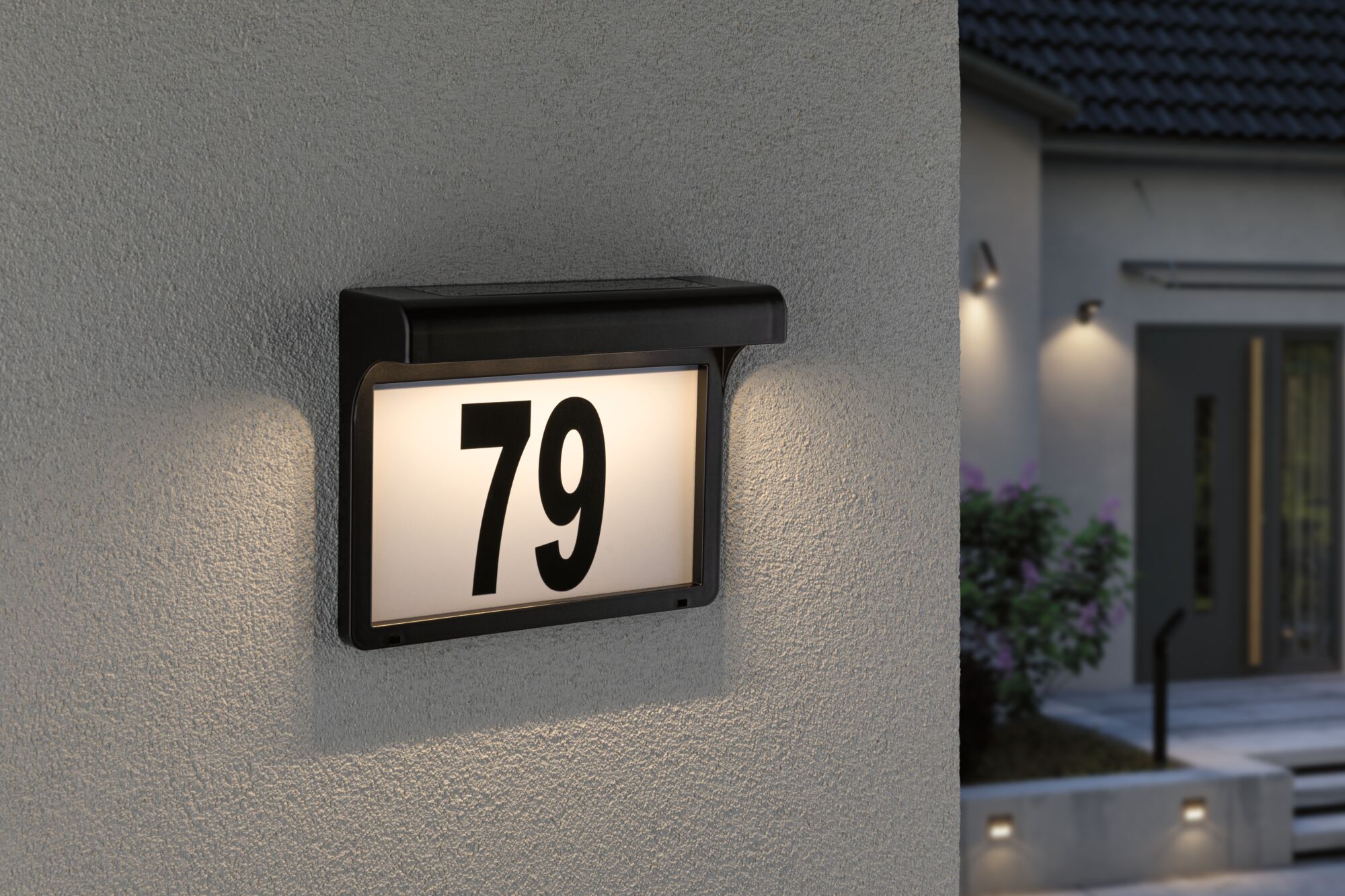Plaque de numéro de maison LED noire avec numéro 79 éclairé sur mur extérieur gris au crépuscule