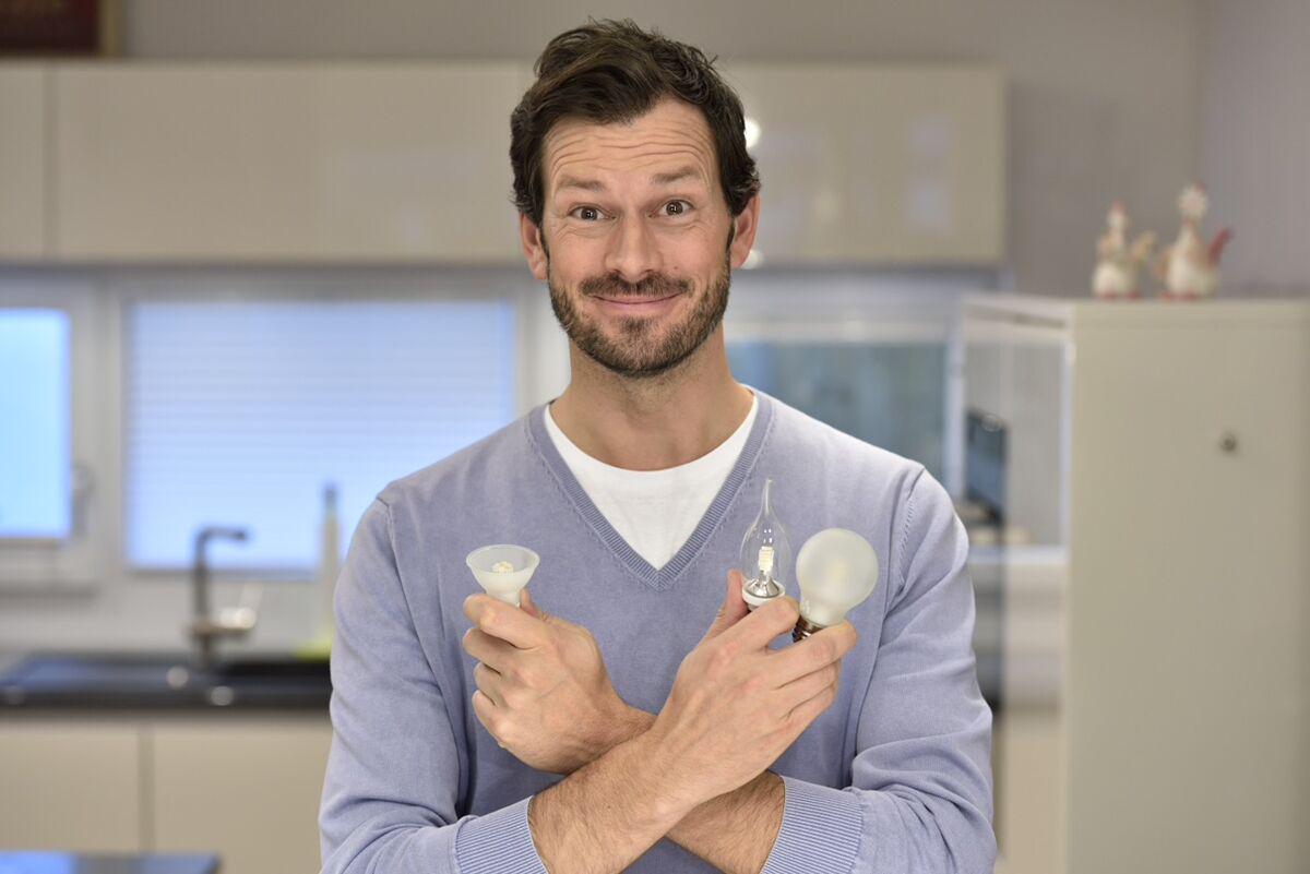 Man in purple sweater holding various light bulbs and LED lamps in modern kitchen, lighting options.